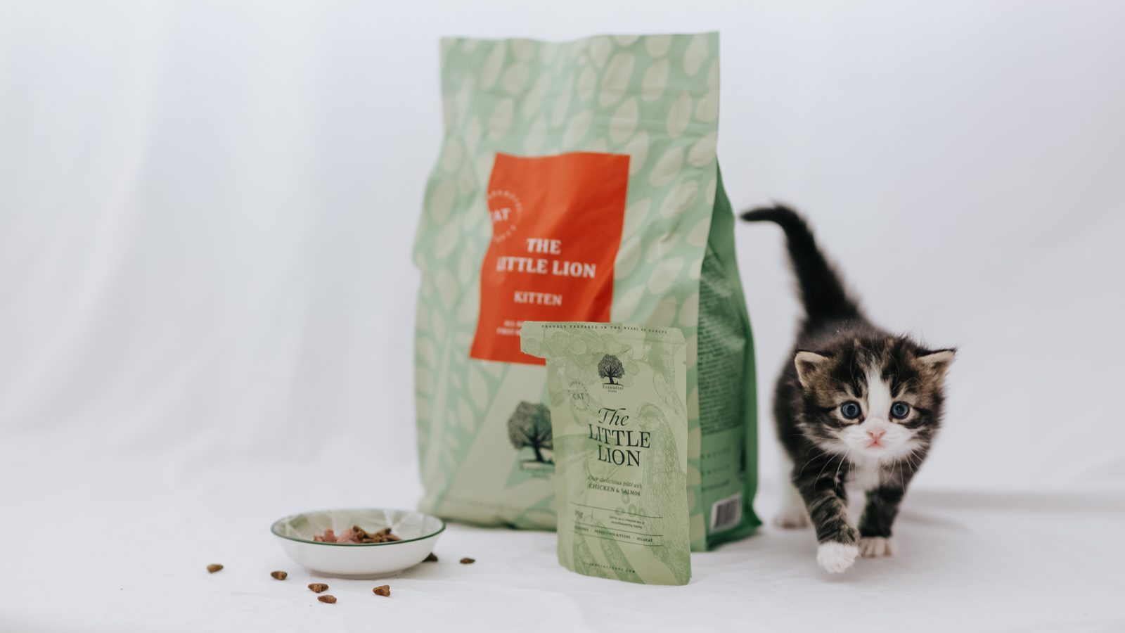 Kitten loopt langs een zak The Little Lion en een zakje Little Lion Paté van Essential Foods, met een bakje voer